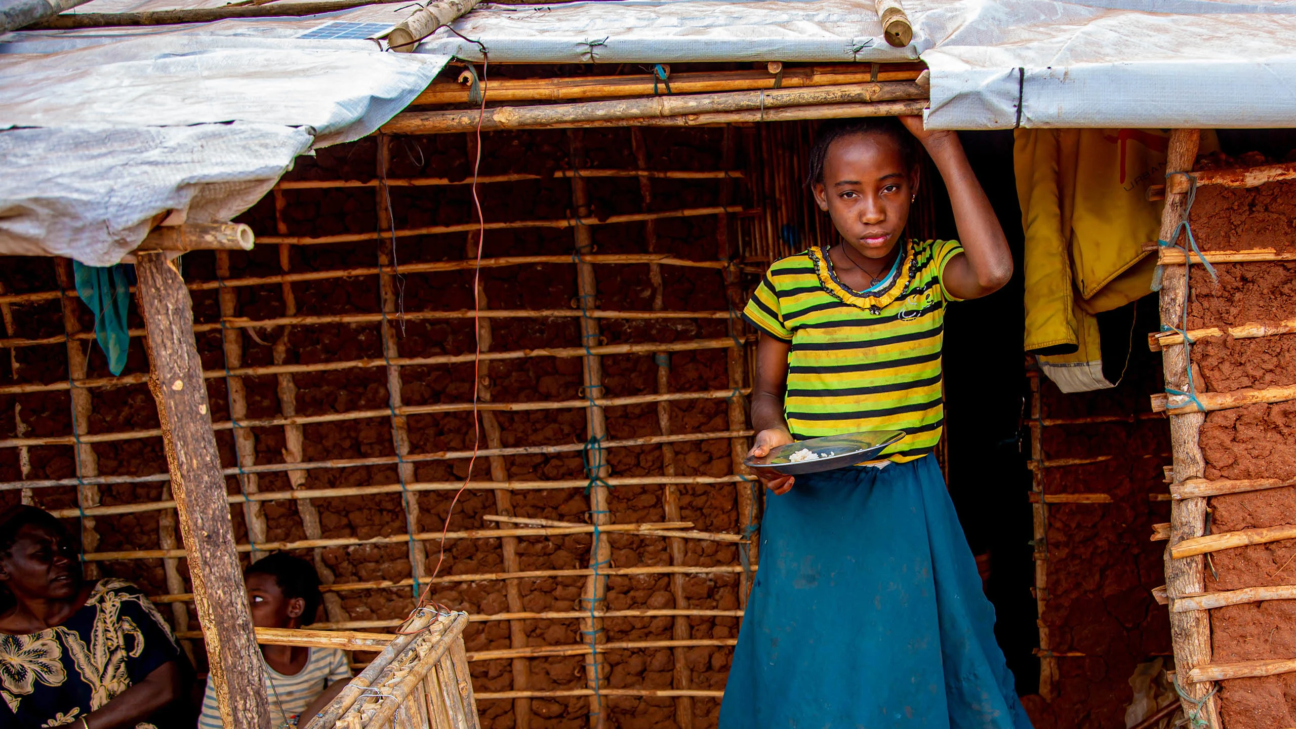 Girl in yellow shirt stands in front of her mud house with an almost empty plate of food.
