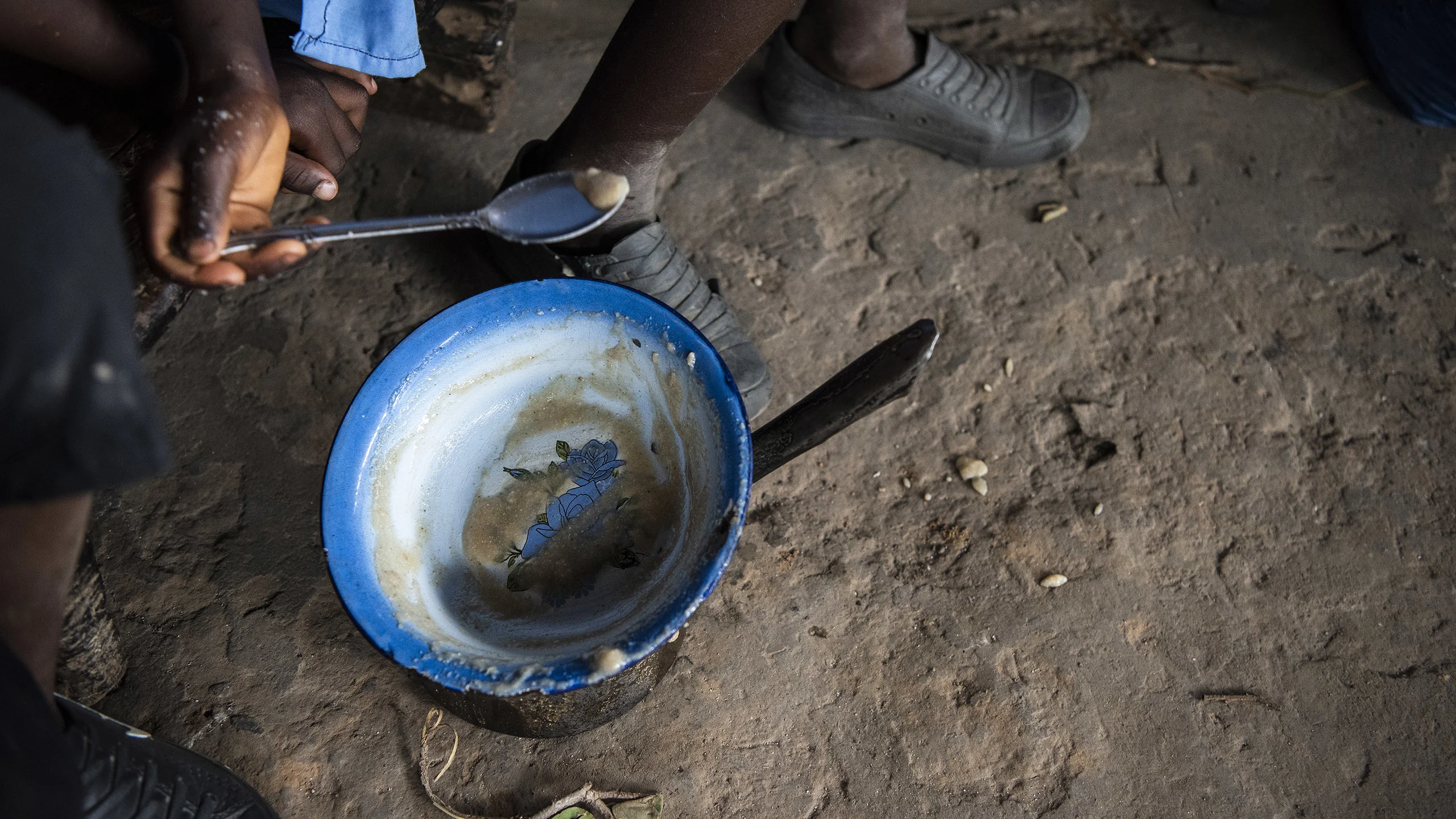 A boy spoons out food from a blue and white bowl on a dirt floor.