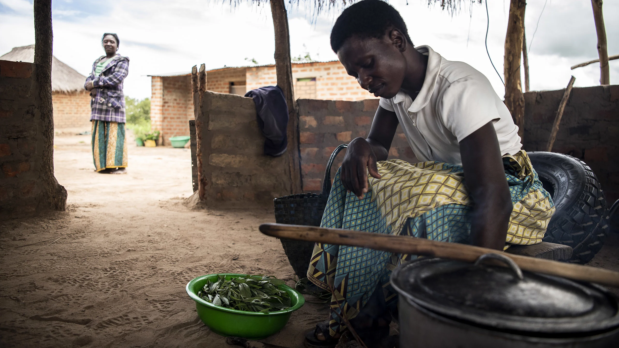 A woman prepares a bowl of plant leaves in her dirt floored kitchen.