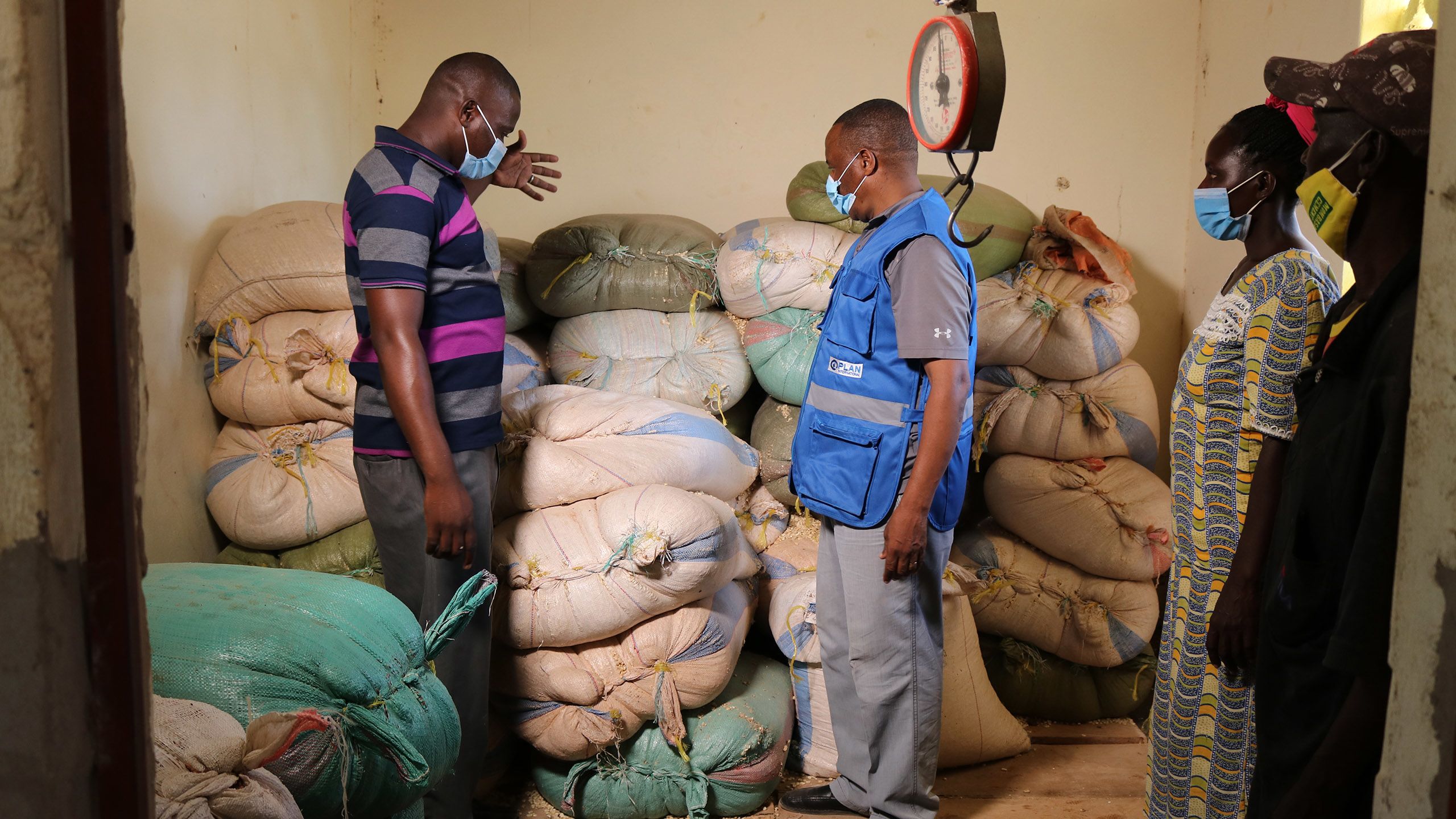 Plan International staff and community memebers stand in front of bags of grain in a distribution centre