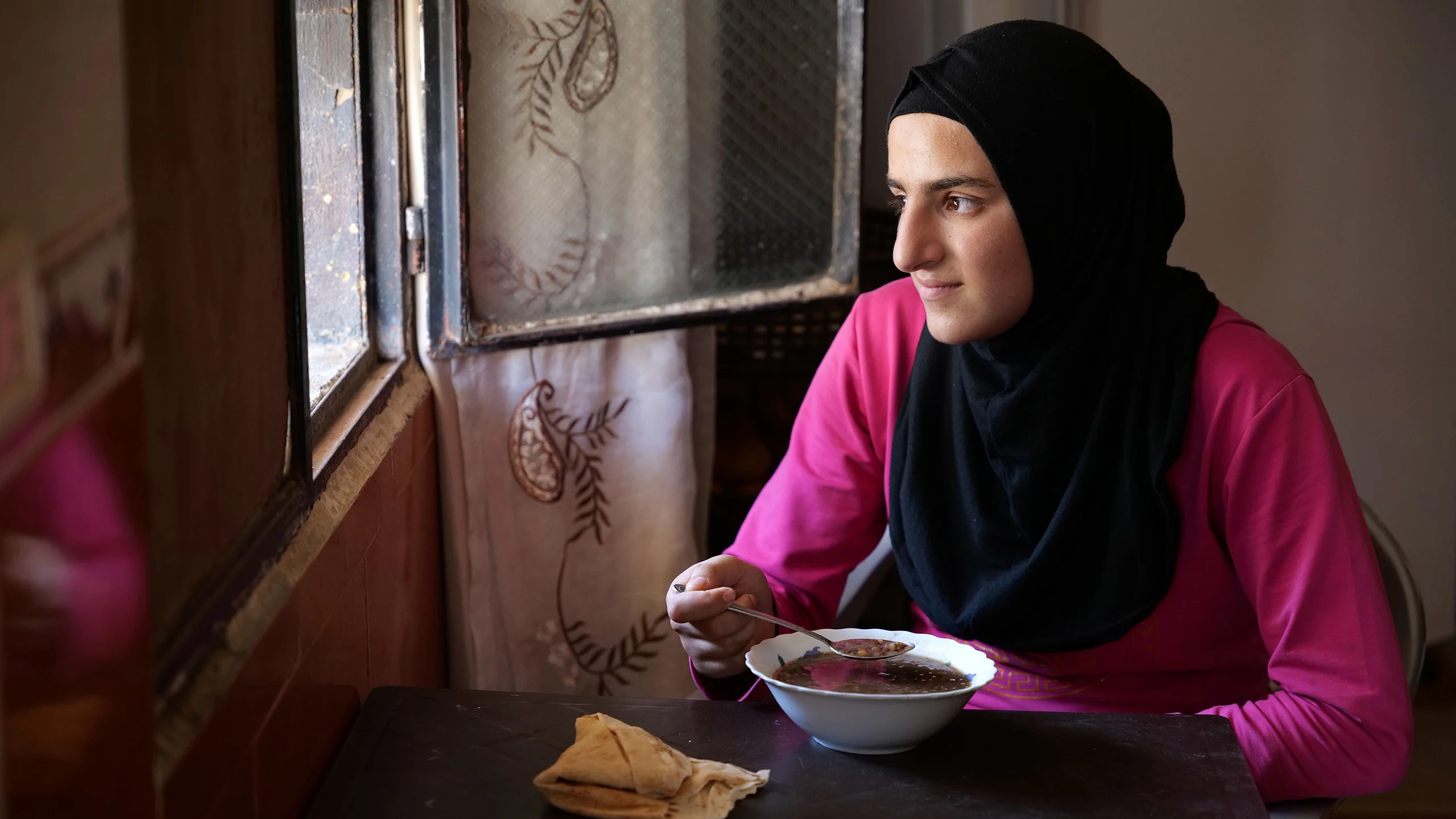 Young woman in a pink shirt and black Hijab sits next to a window with a full bowl of soup.