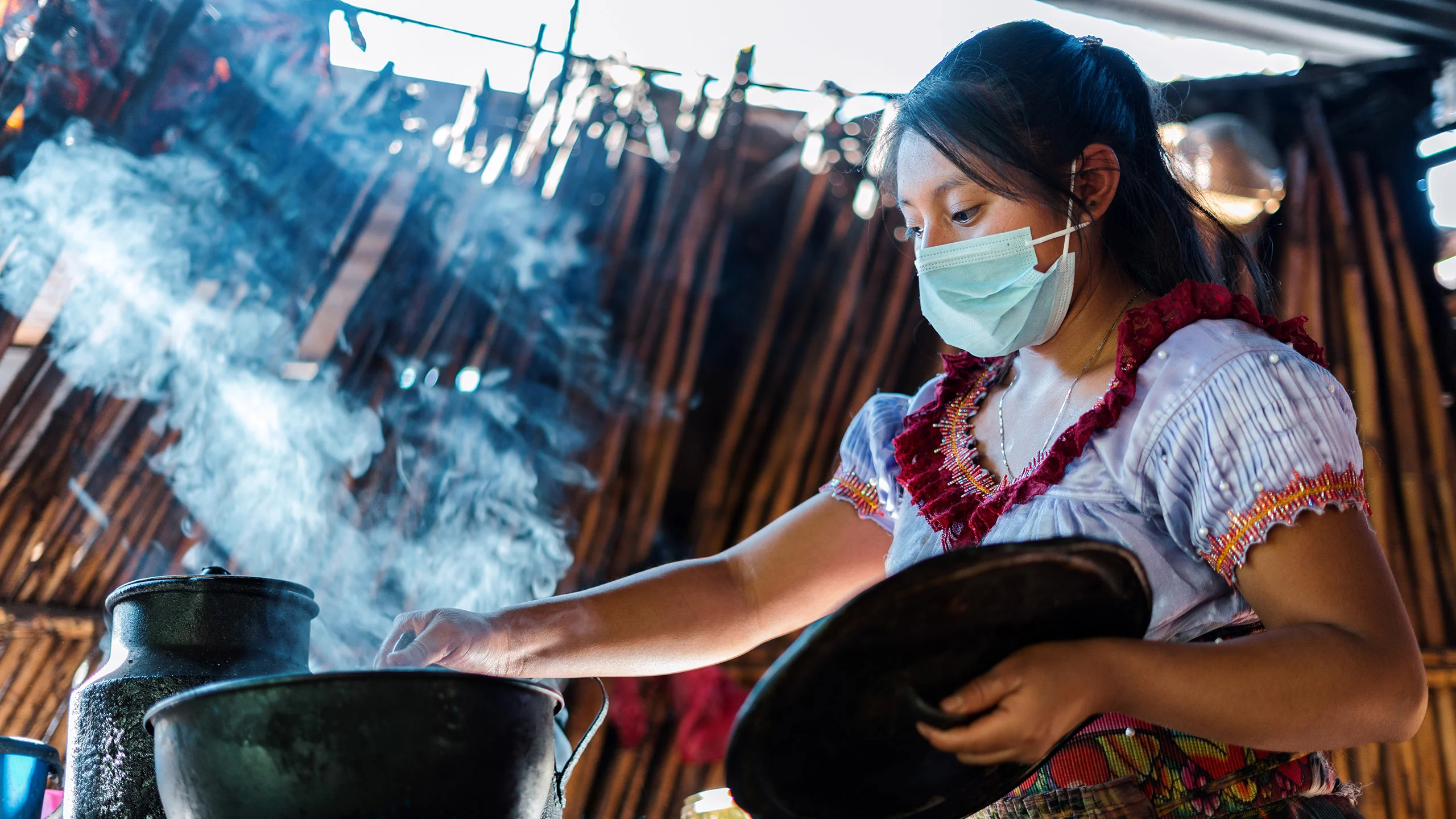 Young woman wearing a mask stirs a pot of steaming food.